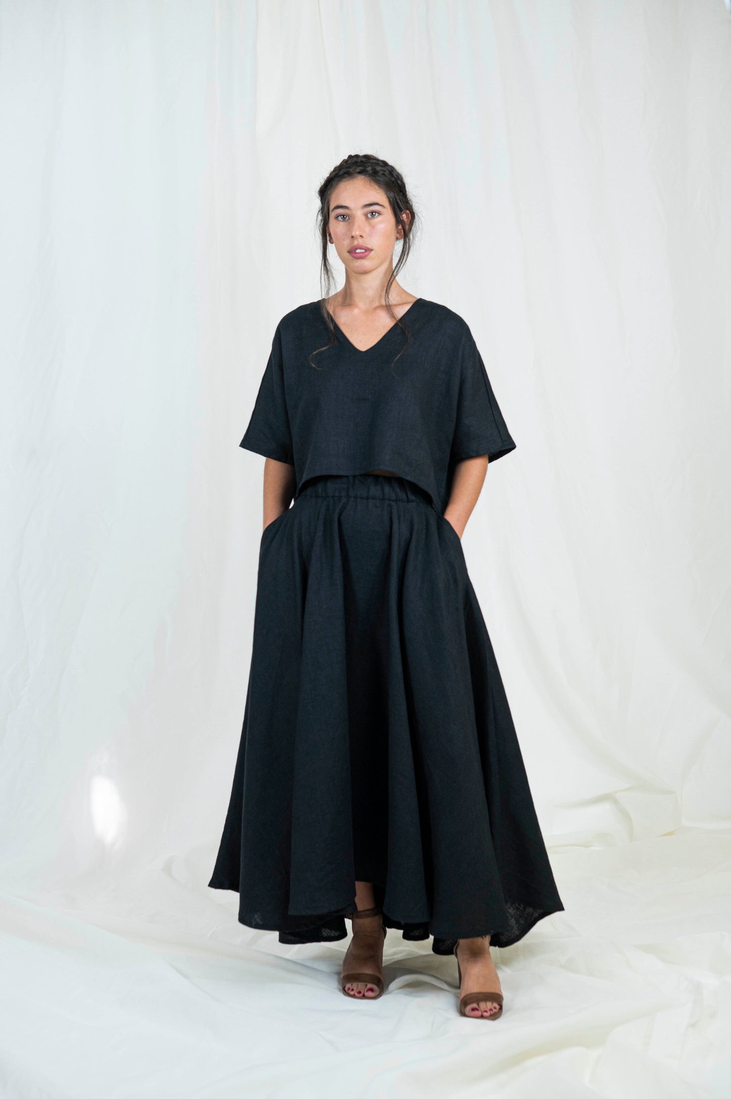 Woman wearing black linen boxy top, softly cropped with a kimono sleeve and v-neck. A full circle skirt with pockets against a white backdrop . We see her facing forward.