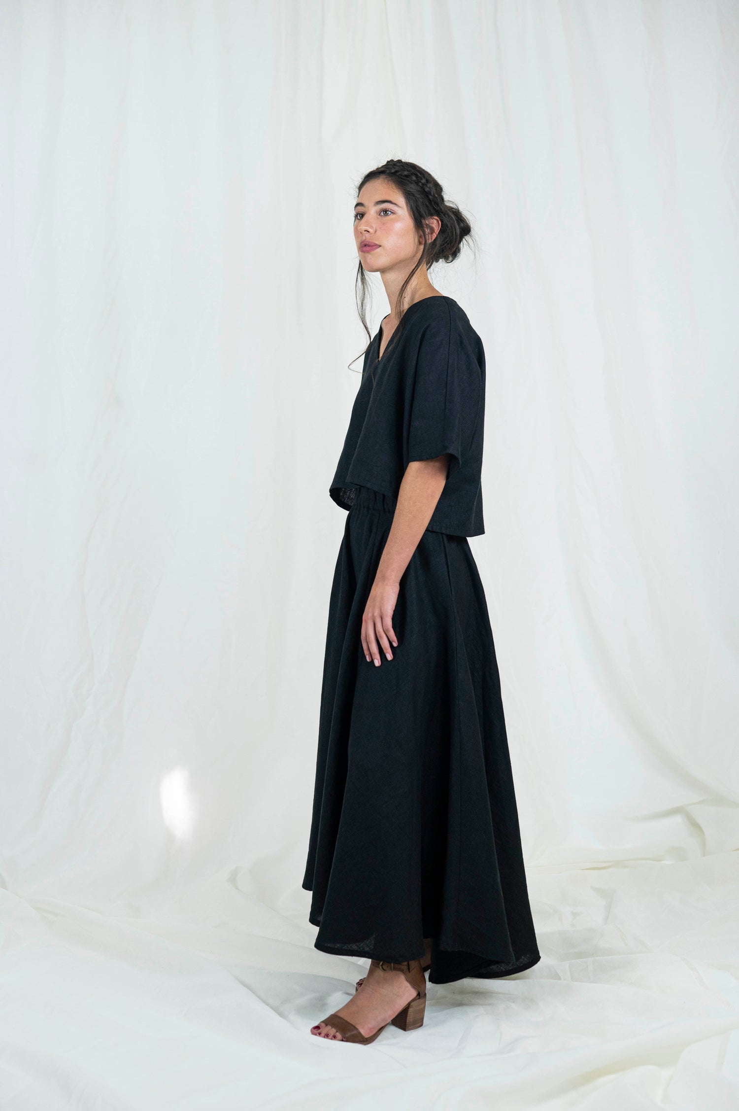 Woman wearing black linen boxy top, softly cropped with a kimono sleeve and v-neck. A full circle skirt with pockets against a white backdrop . We see her standing from the side gazing outward.