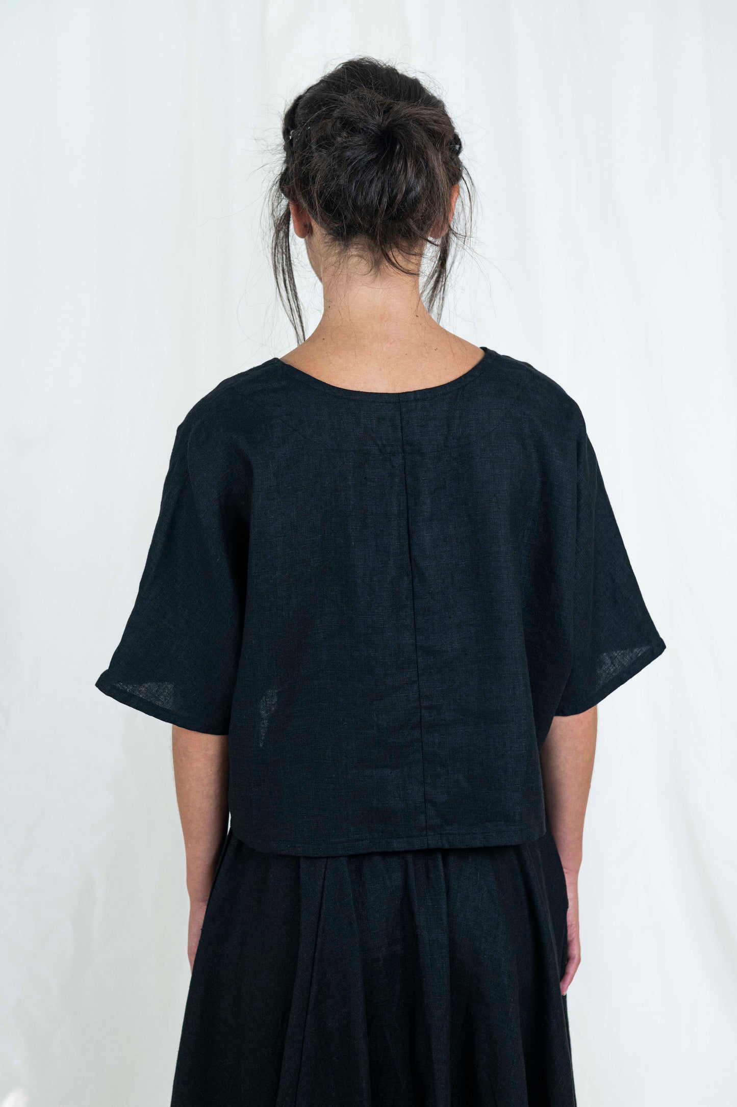 Woman wearing black linen boxy top, softly cropped with a kimono sleeve and v-neck. A full circle skirt with pockets against a white backdrop . We see her back as she faces forward, showing the center back seaming of the top. 