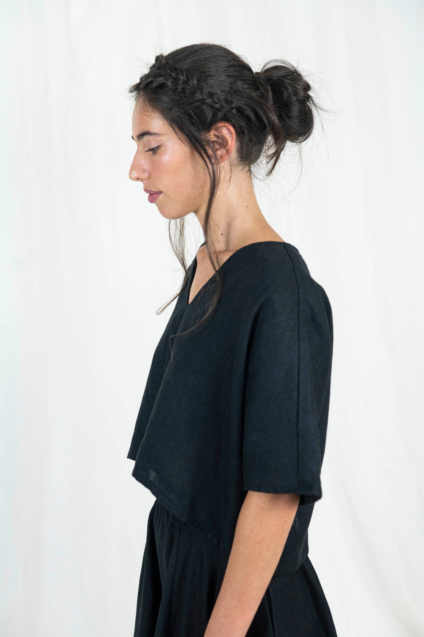 Zoomed in view of woman wearing black linen boxy top, softly cropped with a kimono sleeve and v-neck. A full circle skirt with pockets against a white backdrop . We see her standing from the side gazing downward.ward.