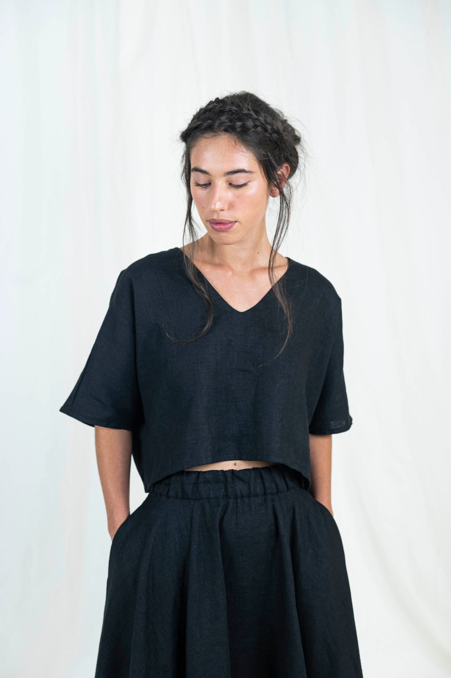 Woman wearing black linen boxy top, softly cropped with a kimono sleeve and v-neck. A full circle skirt with pockets against a white backdrop . We see her standing from the side gazing downward.