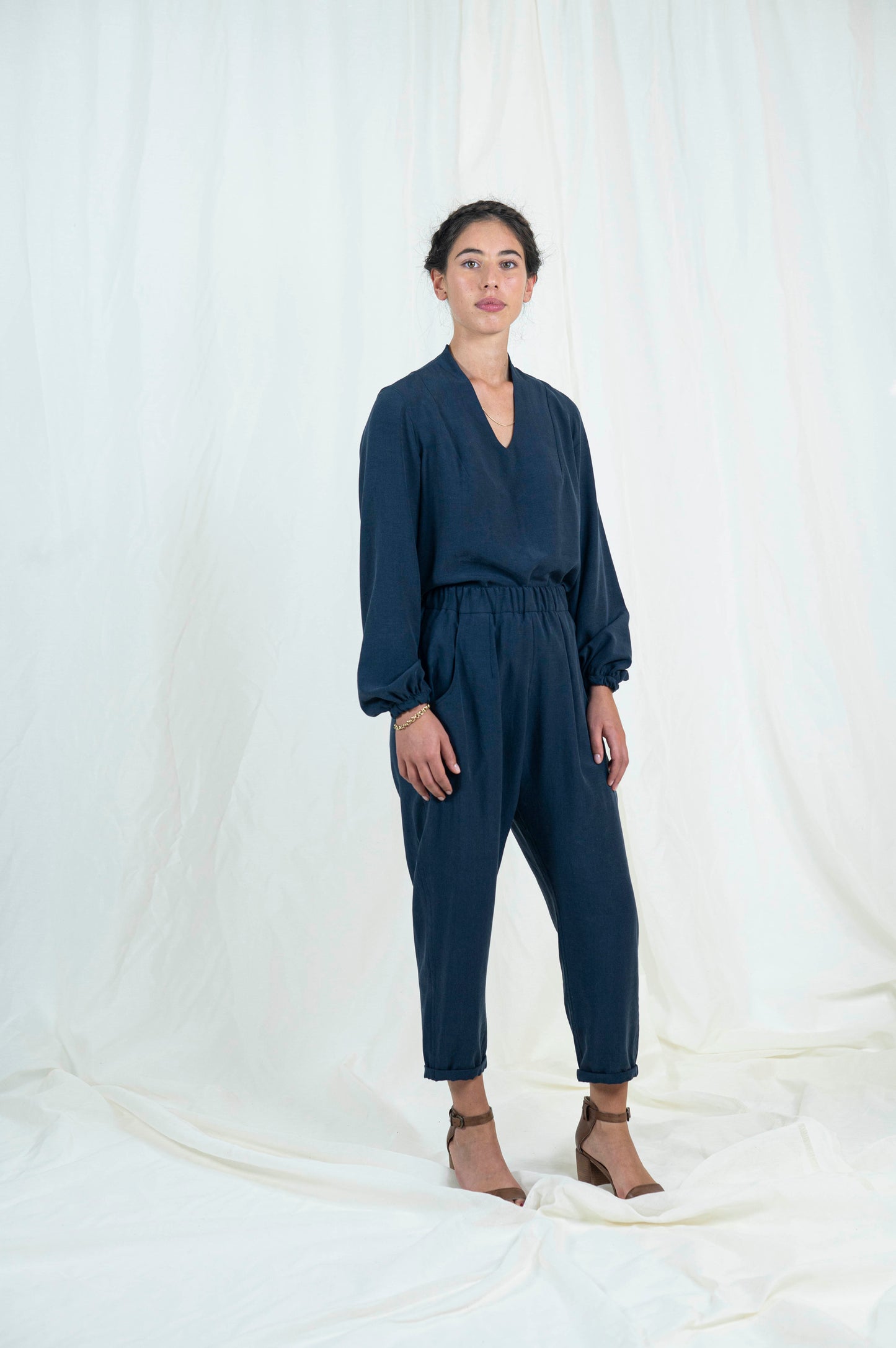 Slightly skewed front view of woman wearing navy blue relaxed pants with pockets and cuffed ankle length hem. Blouse with billowing bishop sleeves cuffed at wrist a high back neck and easy modest v-neckline.