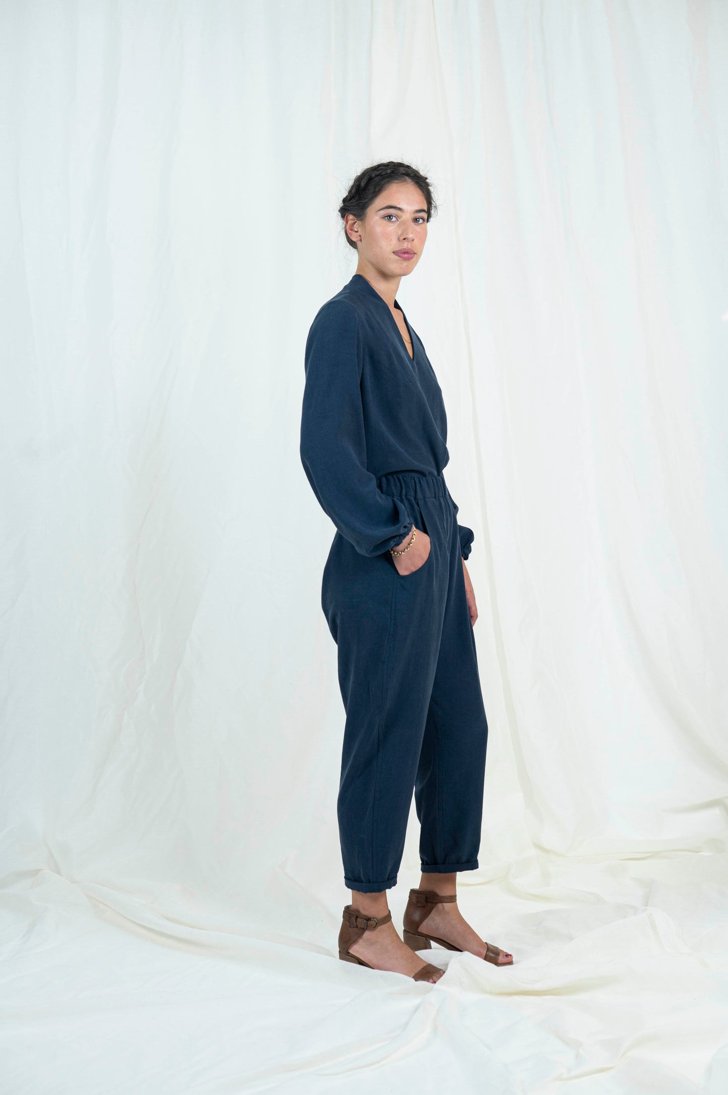 Woman wearing navy blue relaxed pants with pockets and cuffed ankle length hem. Blouse with billowing bishop sleeves cuffed at wrist a high back neck and easy modest v-neckline.