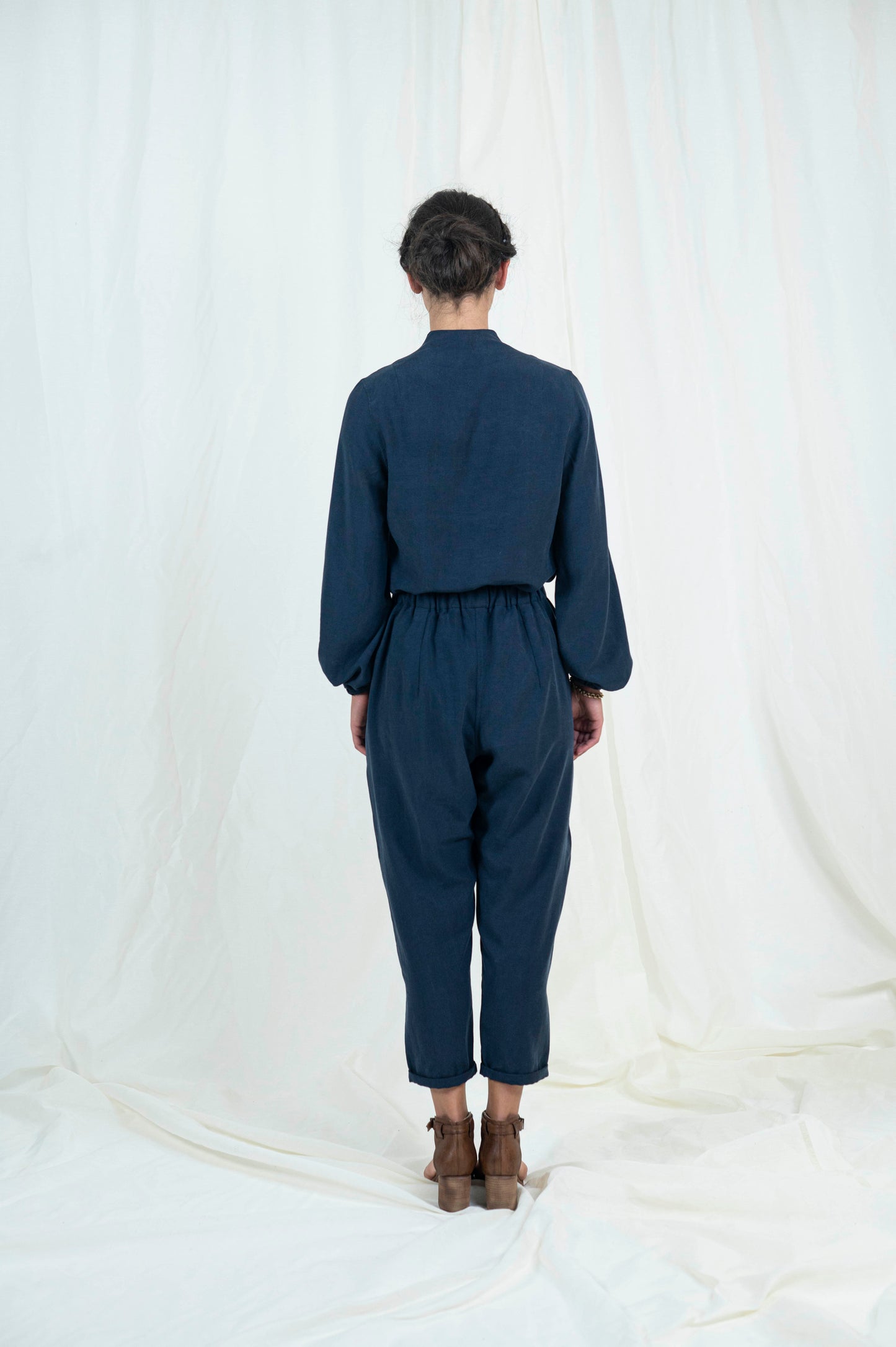 Back view of woman wearing navy blue relaxed pants with pockets and cuffed ankle length hem. Blouse with billowing bishop sleeves cuffed at wrist a high back neck and easy modest v-neckline.