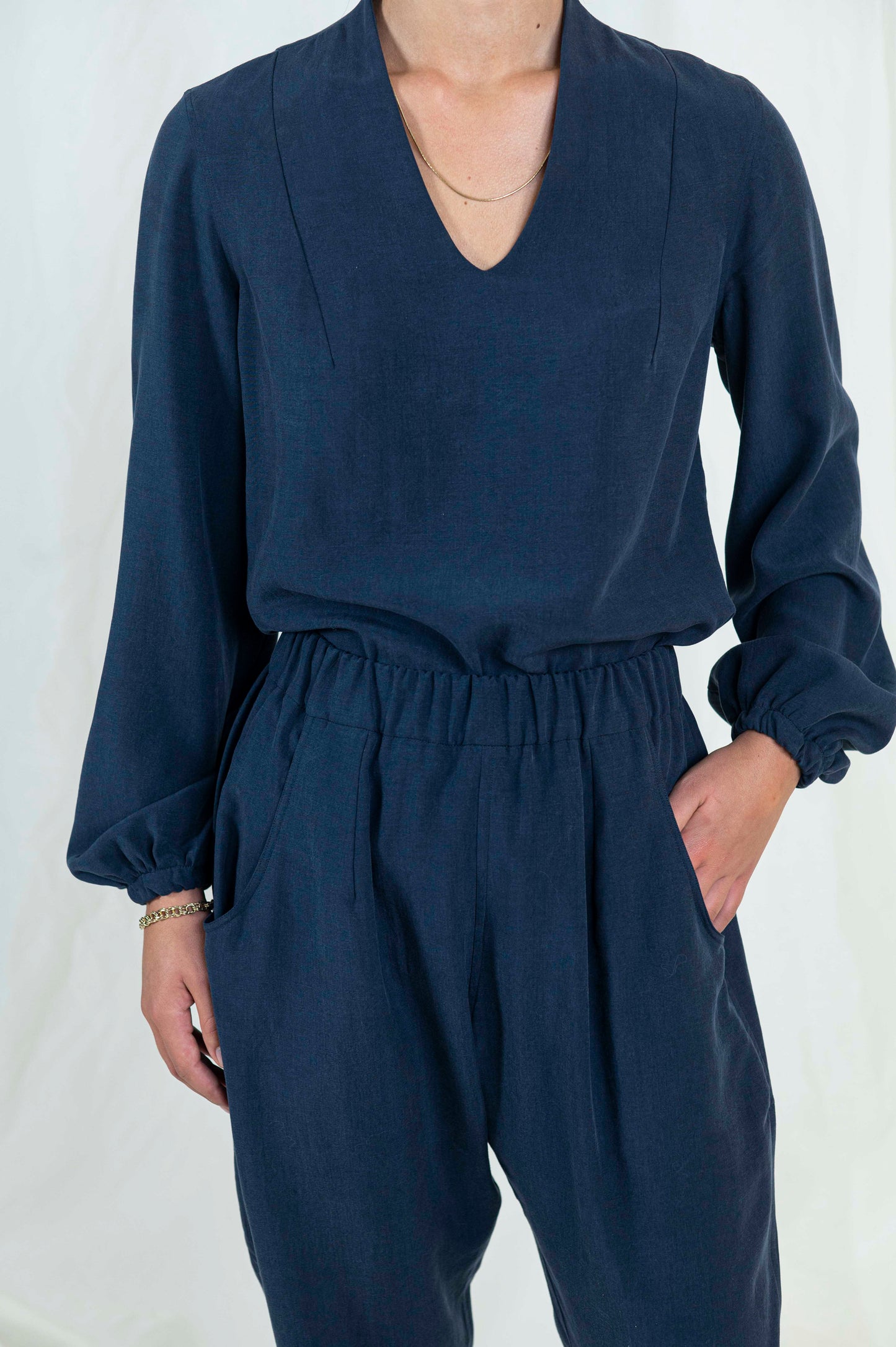 Close up view of woman wearing navy blue relaxed pants with pockets and cuffed ankle length hem. Blouse with billowing bishop sleeves cuffed at wrist a high back neck and easy modest v-neckline. Elastic waistband and dart seams are apparent.