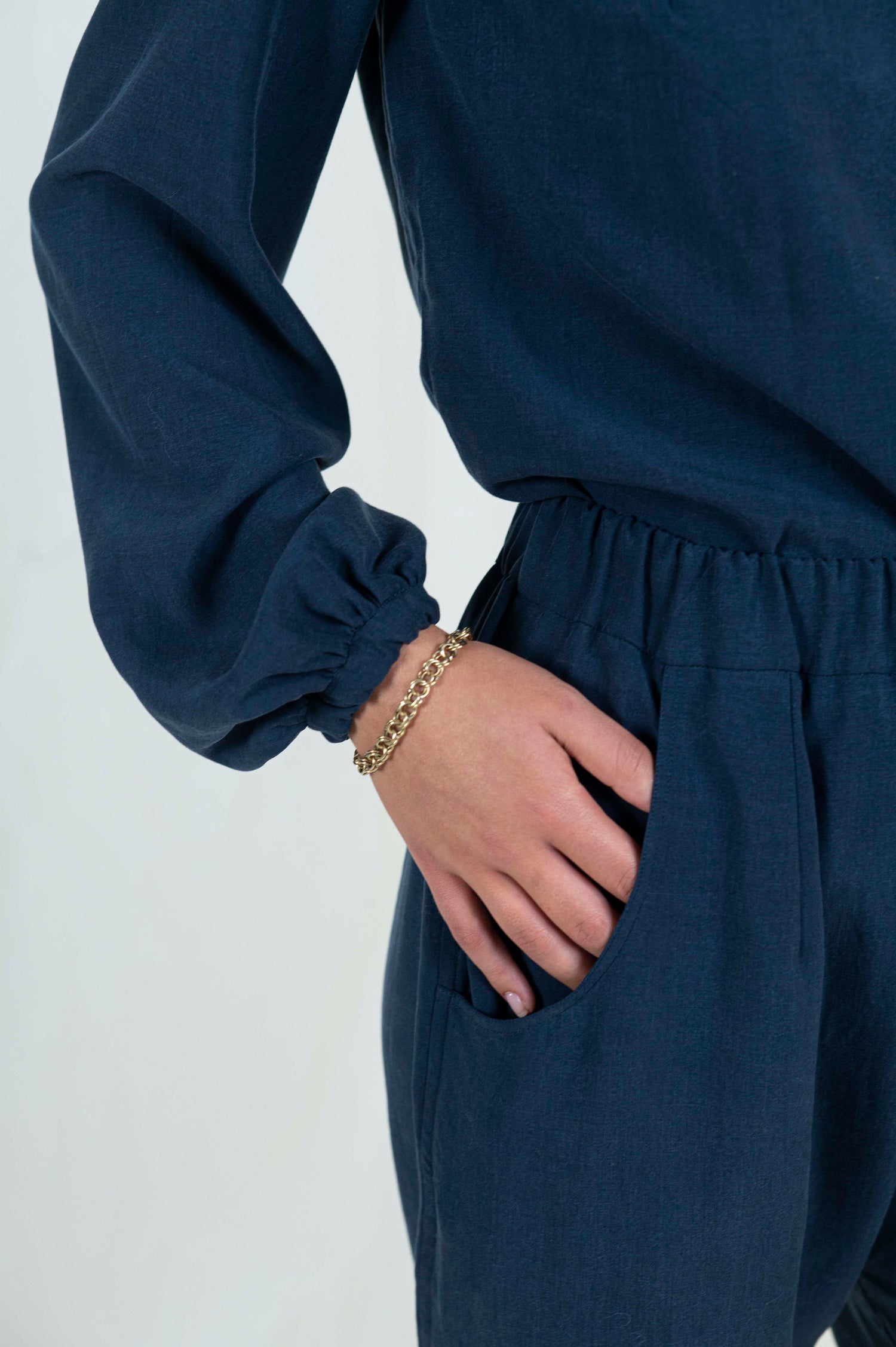 Close-up view of woman's hand in pocket of navy blue relaxed pants with pockets and cuffed ankle length hem. Blouse with billowing bishop sleeves cuffed at wrist a high back neck and easy modest v-neckline.