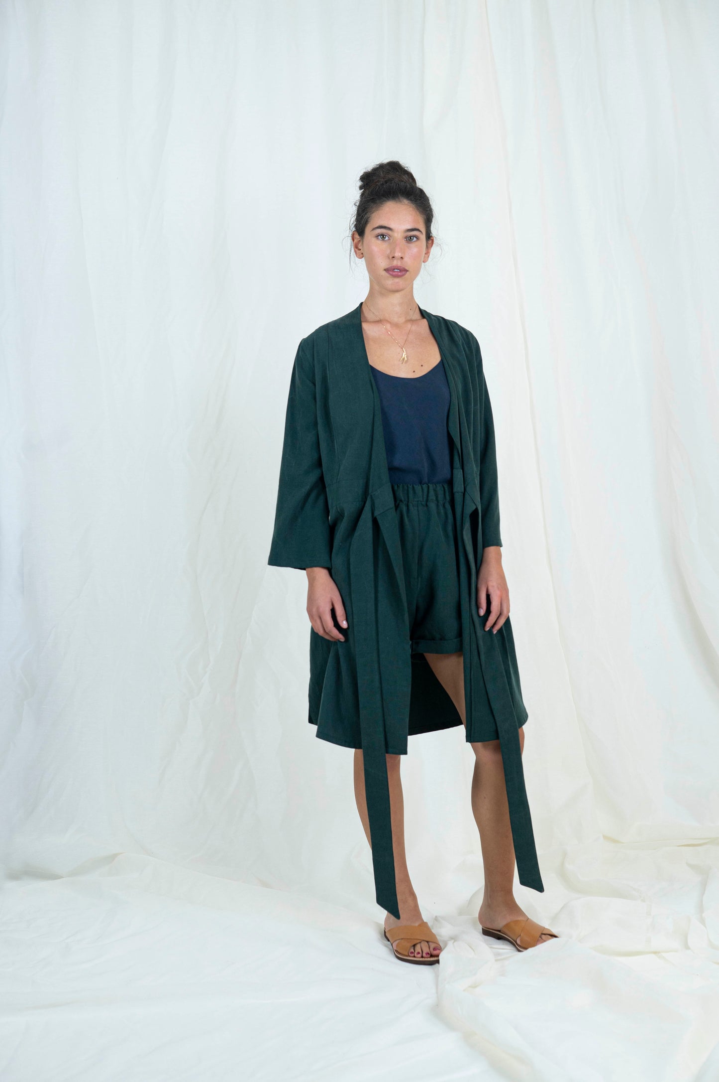 Dark haired woman wearing dark green shorts, navy blue camisole with open dark green robe, front view.