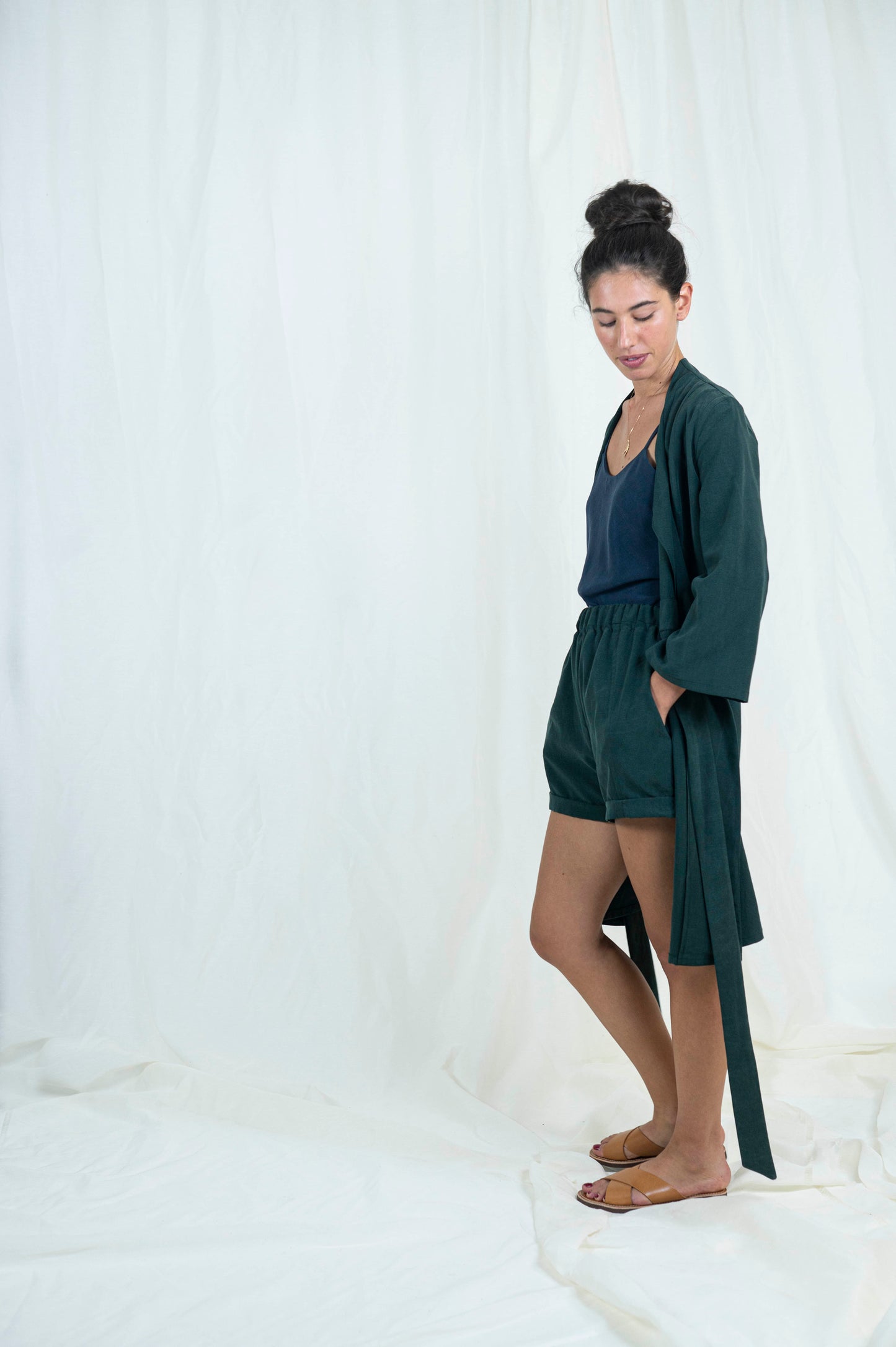 Dark haired woman wearing dark green shorts, navy blue camisole with open dark green robe, from the side gaze is downward.