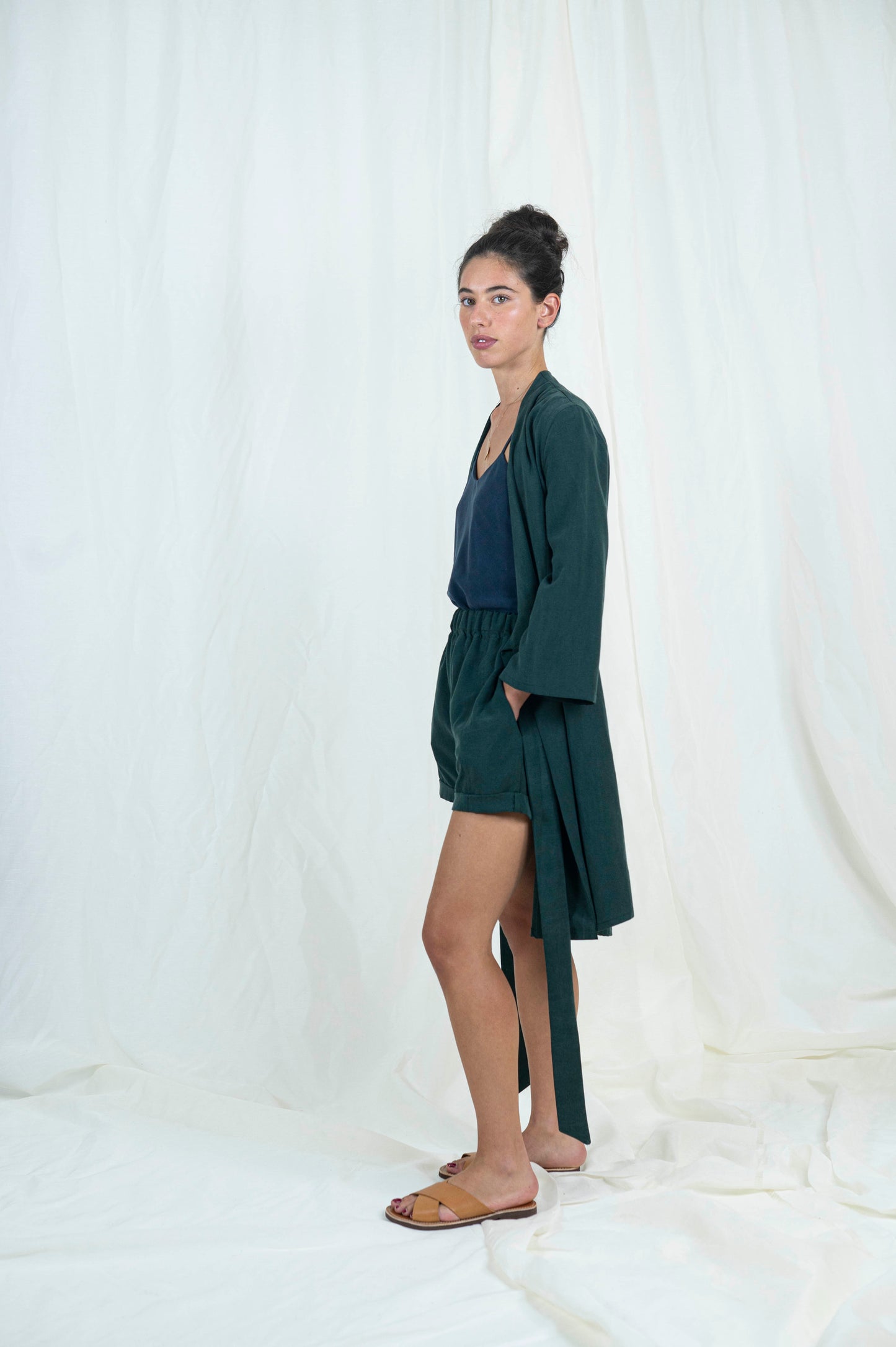 Dark haired woman wearing dark green shorts, navy blue camisole with open dark green robe, from the side.