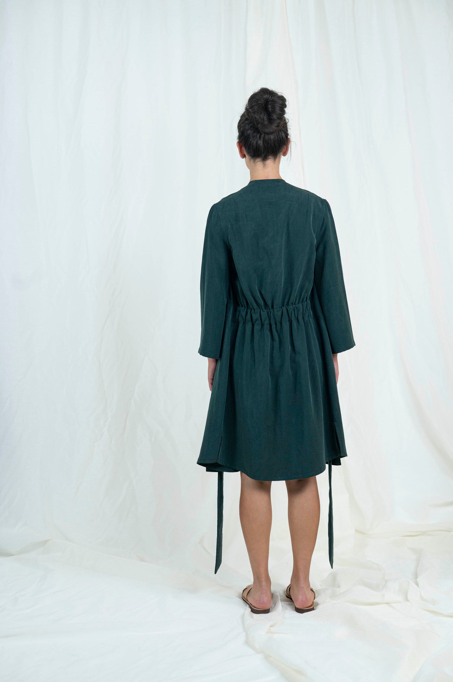 Dark haired woman wearing dark green shorts, navy blue camisole with open dark green robe, viewed from the back.