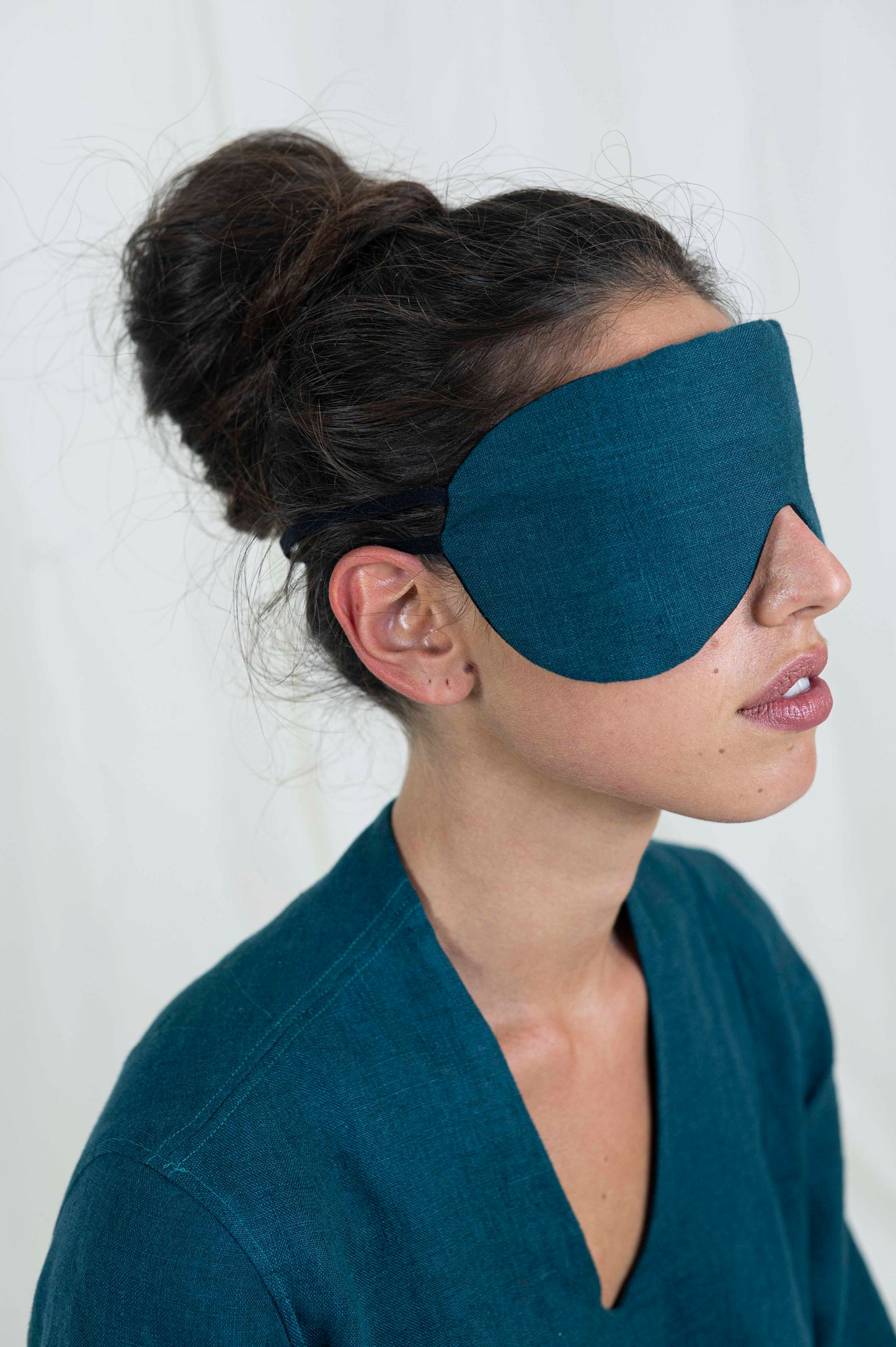 Dark haired woman from the side, eyes covered with sleep mask. With a white background and linen dress.