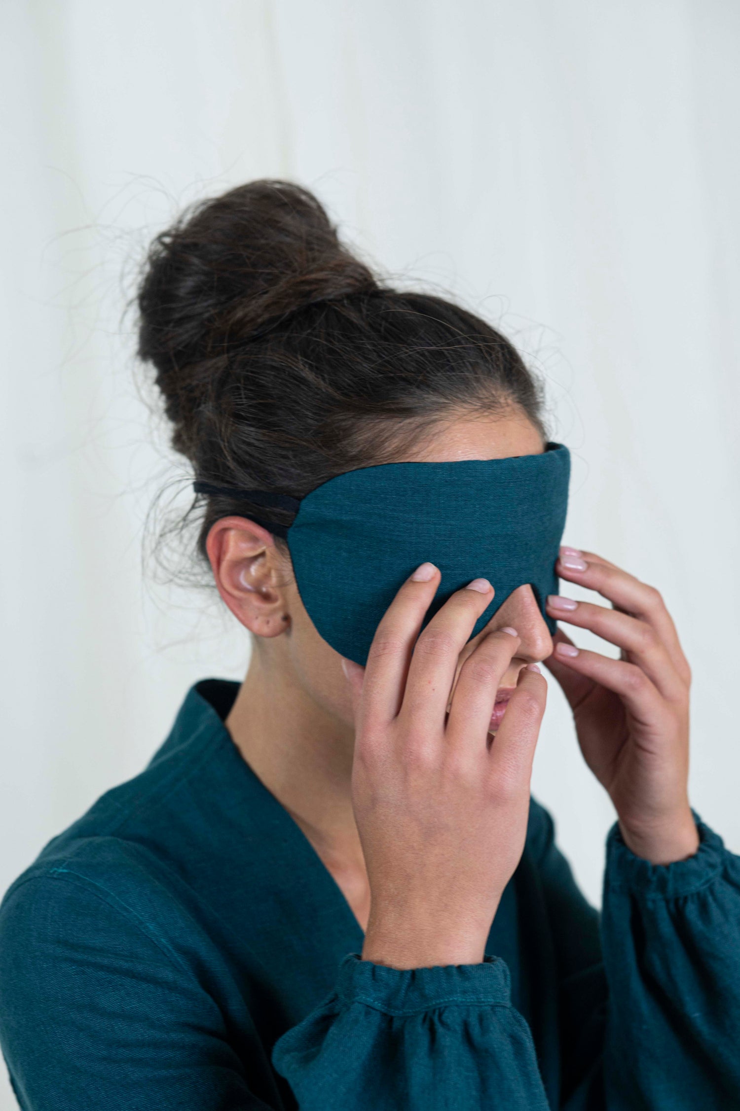 Dark haired woman her hands touching seep mask covering her eyes. With a white background and linen dress.