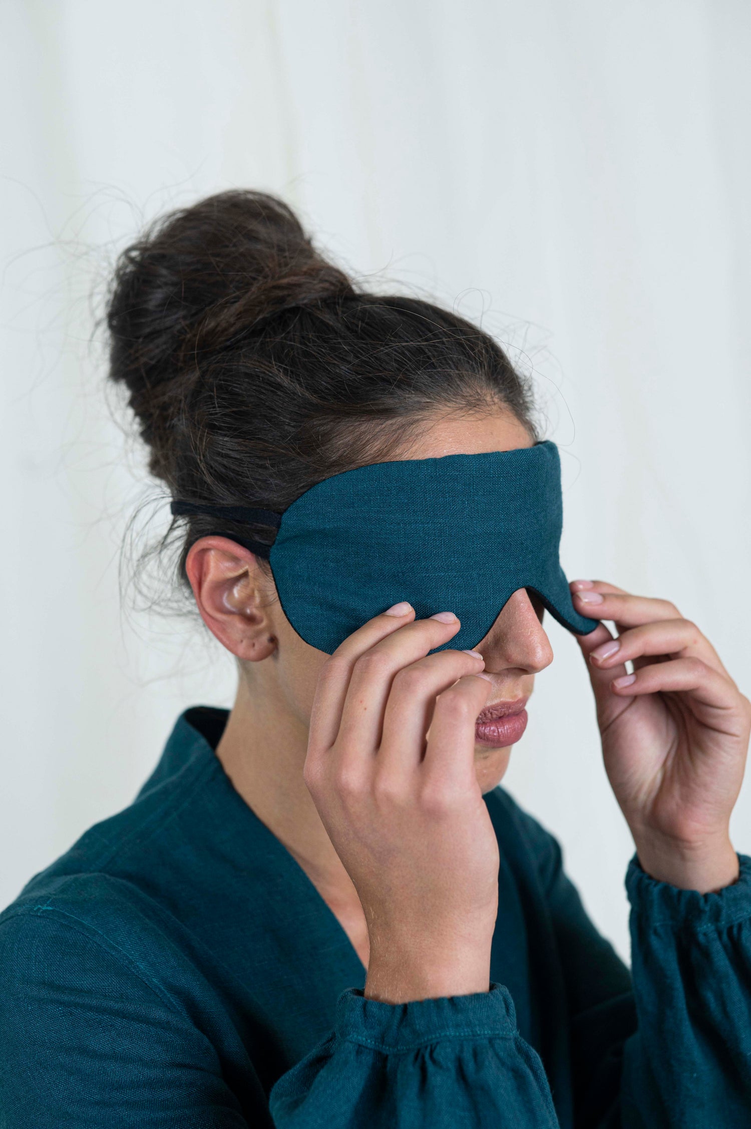 Dark haired woman eyes covered with sleep mask. With a white background and linen dress.