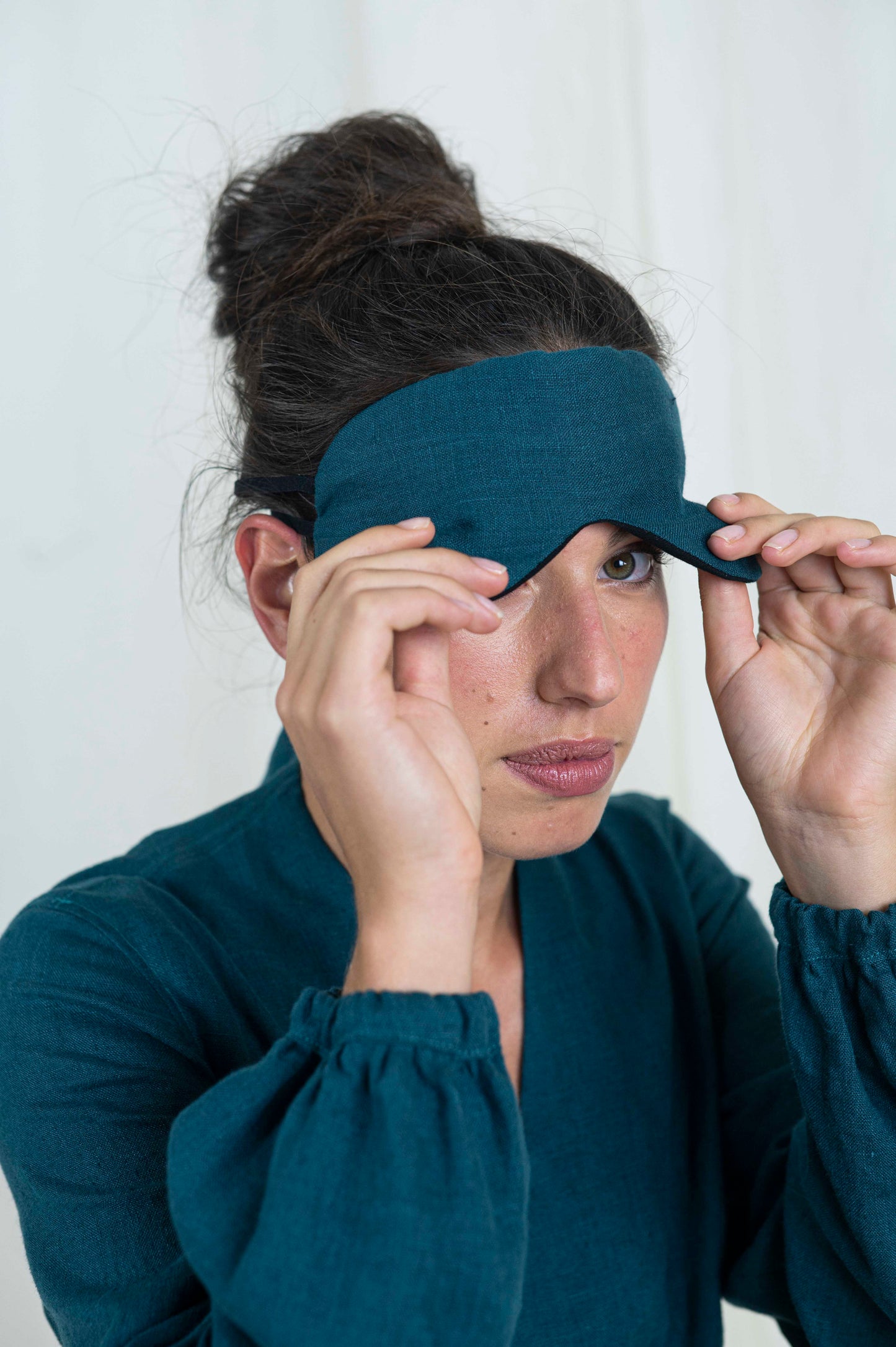 Dark haired woman peaking out from sleep mask. With a white background and linen dress.