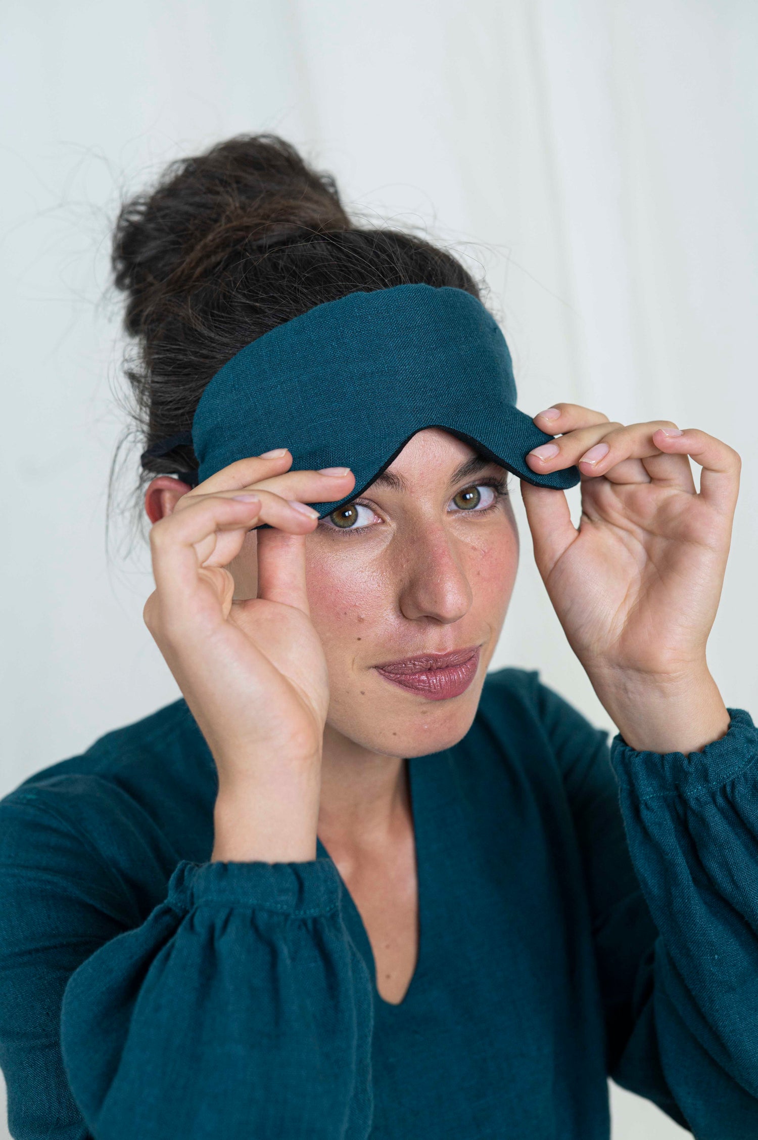 Dark haired woman peaking out from sleep mask. With a white background and linen dress.