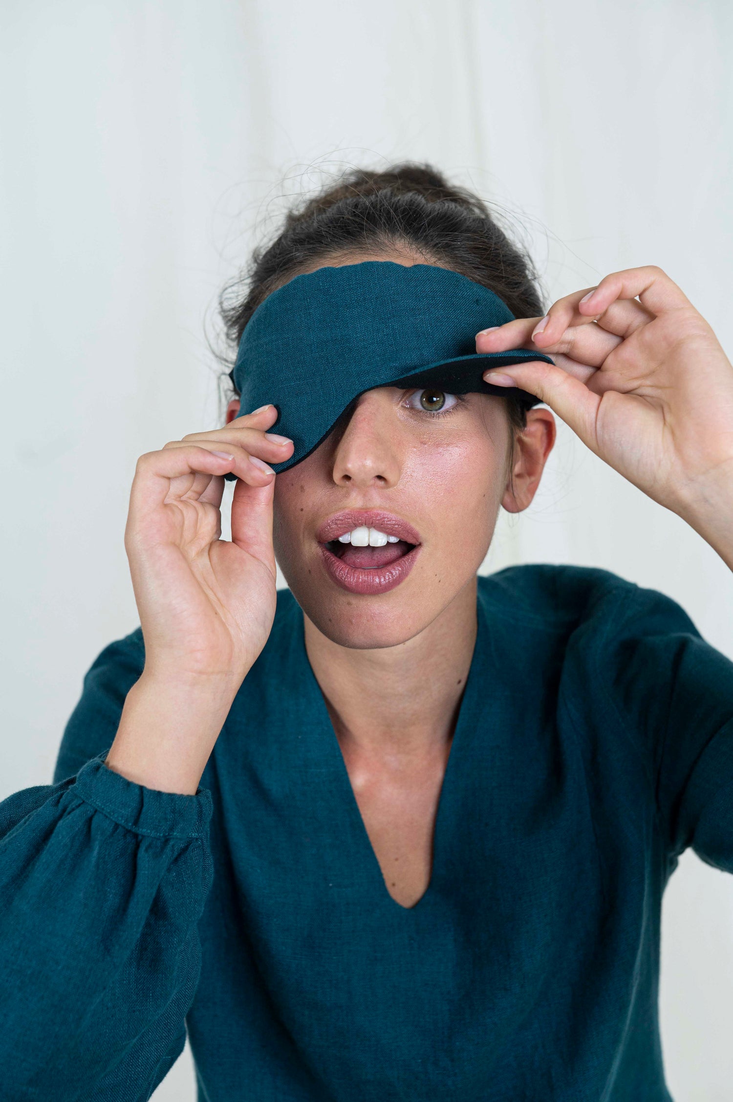 Dark haired woman peaking out from sleep mask. With a white background and linen dress.