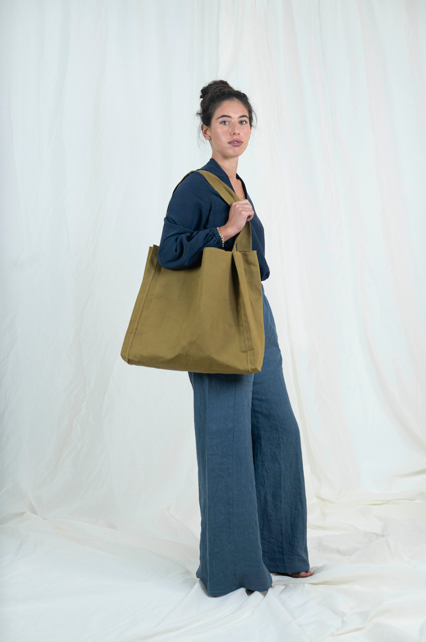 Dark haired woman wearing medium blue wide legged linen pants, dark blue bishop sleeve top and oversized tan canvas tote bag, viewed from the side, gaze is forward. 