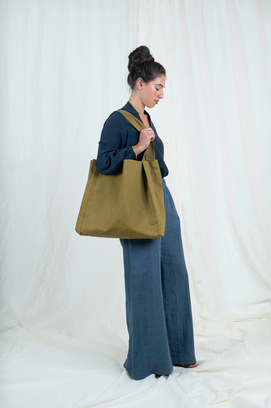 Dark haired woman wearing medium blue wide legged linen pants, dark blue bishop sleeve top and oversized tan canvas tote bag, viewed from the side, gaze is downward. 